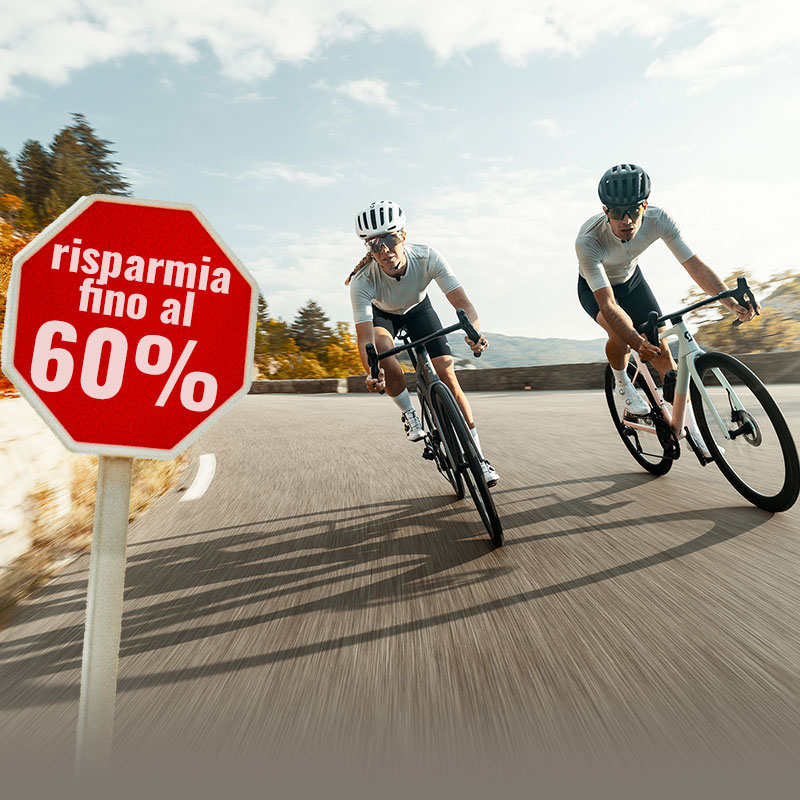Due persone con caschi e abbigliamento ciclismo pedalano su una strada di montagna tortuosa, dove un cartello indica uno sconto fino al 60 percento.