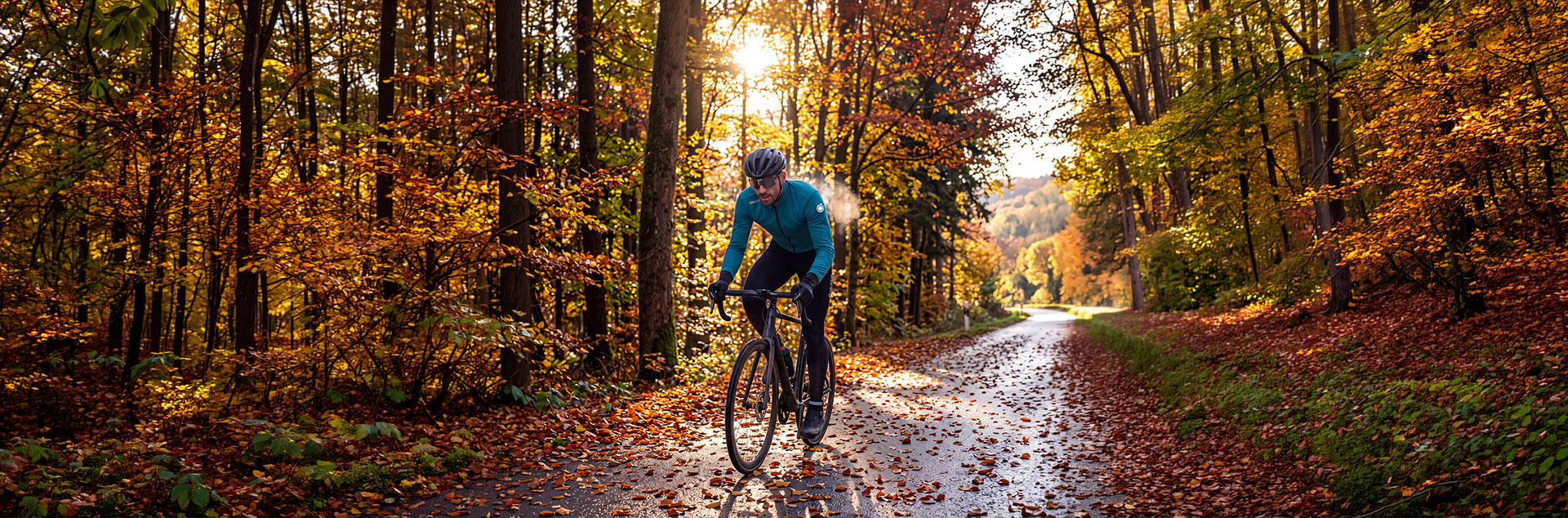 Ein Radfahrer in türkisfarbener Jacke fährt auf einer mit Herbstlaub bedeckten Waldstraße im Licht der Morgensonne.