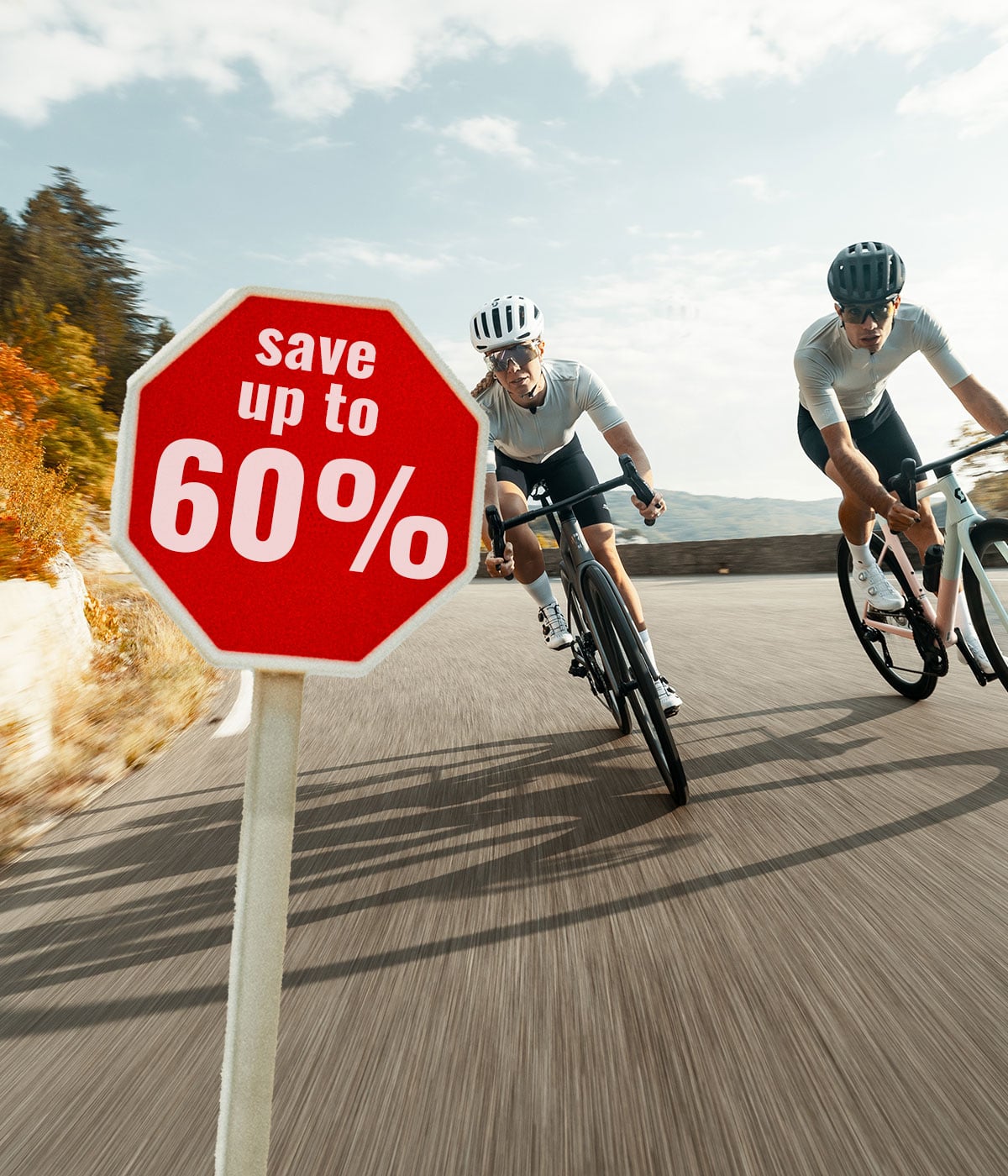 Two people wearing helmets and cycling clothing ride their bikes on a winding mountain road, where a sign indicates a discount of up to 60 percent.