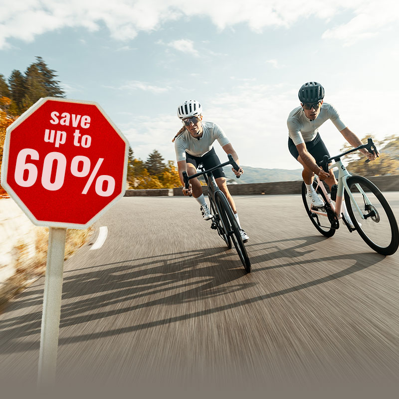 Two people wearing helmets and cycling clothing ride their bikes on a winding mountain road, where a sign indicates a discount of up to 60 percent.