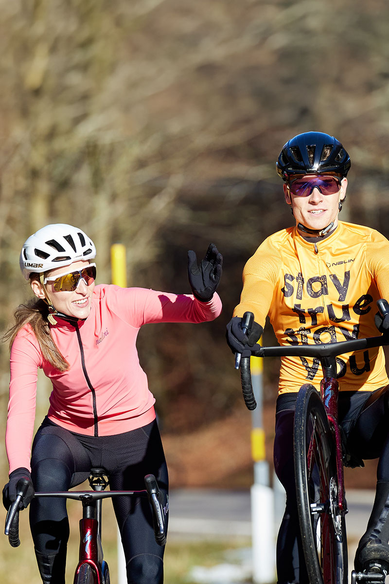 Eine Frau in pinkfarbenem Trikot winkt fröhlich, während sie neben einem Mann in gelbem „Stay True Stay You“-Trikot auf einer sonnigen Herbststraße radelt.