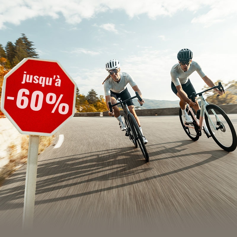 Deux personnes portant des casques et des vêtements cyclistes roulent à vélo sur une route de montagne sinueuse, où un panneau indique une réduction allant jusqu'à 60 %.